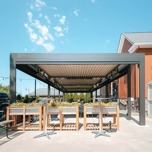 Outdoor dining area featuring a modern metal pergola with retractable roof over wooden tables filled with planters, set against a clear blue sky. The design adds an inviting ambiance to the restaurant's exterior.