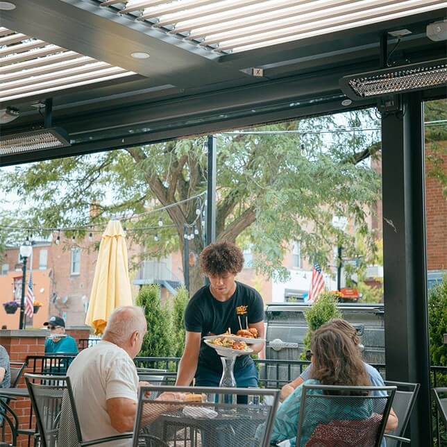 A young waiter serves food to an elderly couple at an outdoor restaurant patio, featuring modern furnishings and open roofing with a view of a quiet urban street.