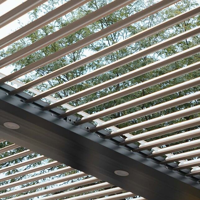 A modern architectural detail showing a series of evenly spaced wooden slats on a metal framework, looking up the structure with a backdrop of green trees and sky, highlighting contemporary design and natural integration.