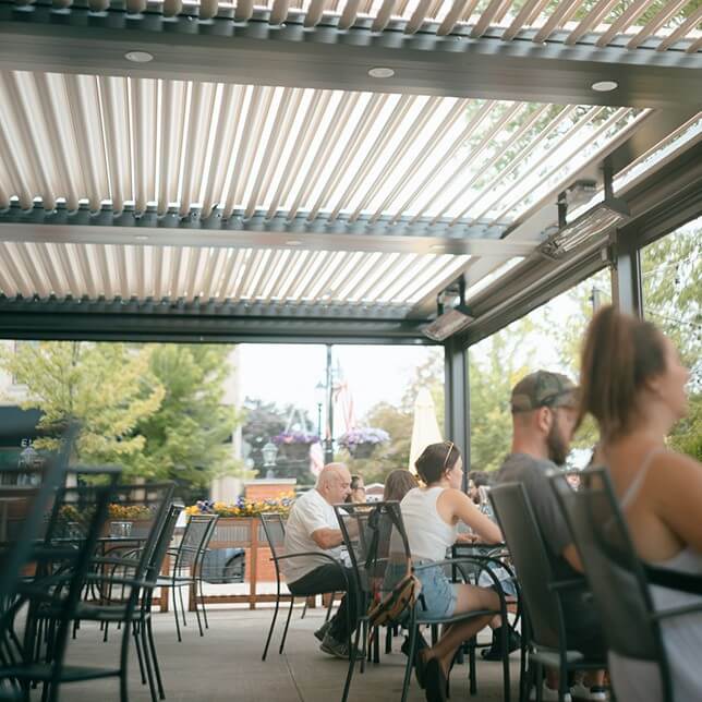 People dining at an outdoor patio area with modern retractable roof, enjoying a sunny day in a relaxed, airy environment surrounded by greenery and city views.