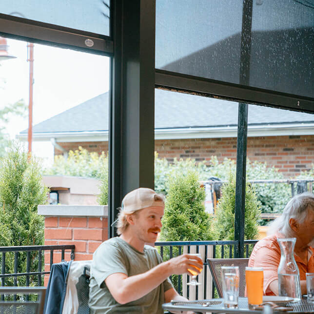 A young man in a casual t-shirt and baseball cap enjoys a glass of beer at an outdoor restaurant patio, featuring green plants and an urban backdrop.
