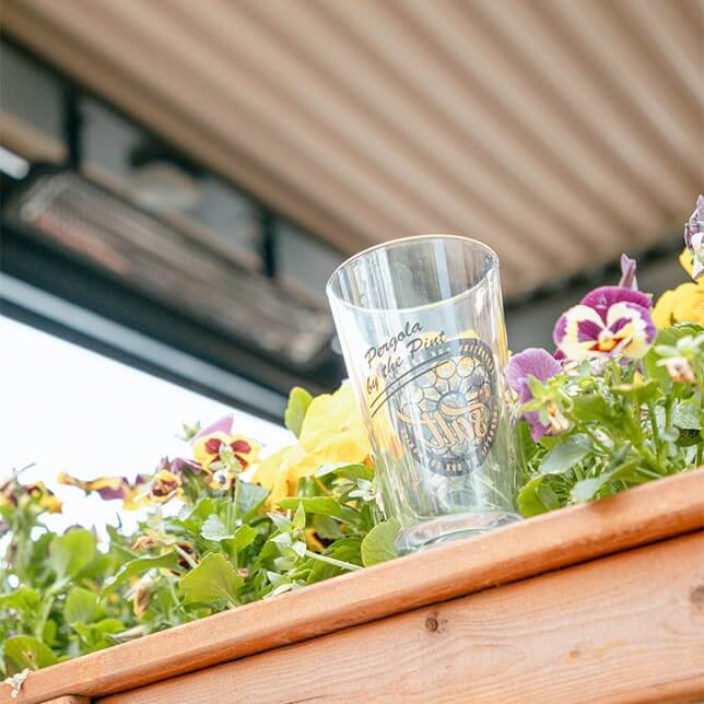 A clear glass featuring decorative lettering stands amidst vibrant pansies in a wooden planter, offering a casual, outdoor ambiance.