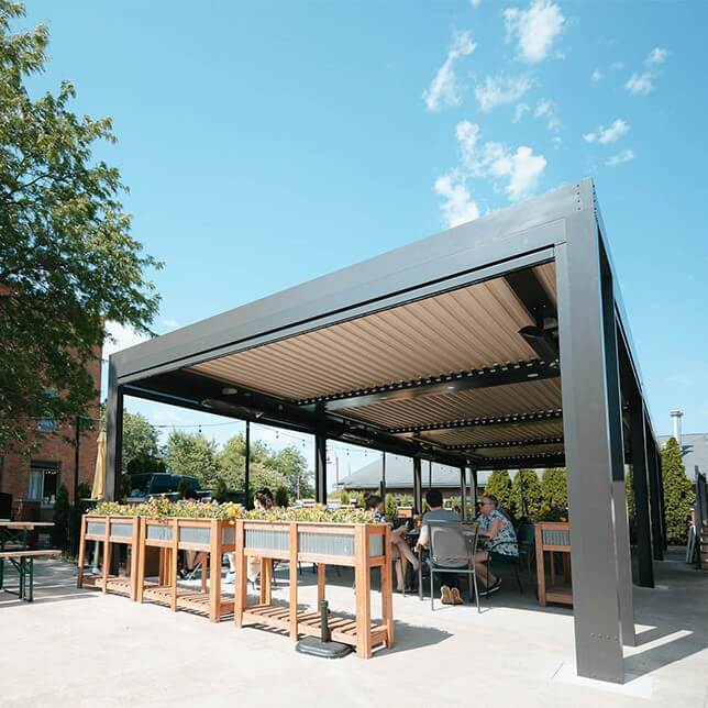 Outdoor seating at a modern restaurant featuring a large, shaded pergola and wooden dining tables surrounded by lush planters, with patrons enjoying a sunny day.