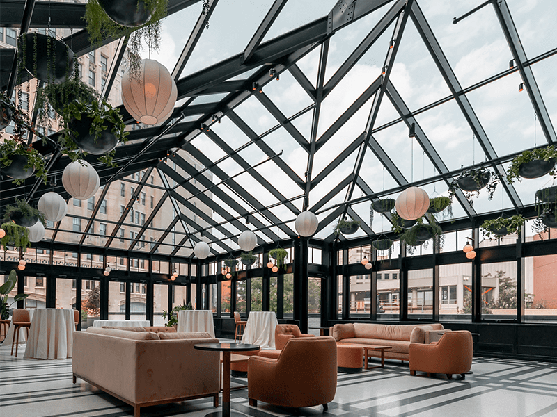 Modern cafe exterior with large glass windows and wooden accents, featuring an outdoor seating area with multiple tables and chairs on a patio surrounded by a landscaped garden.