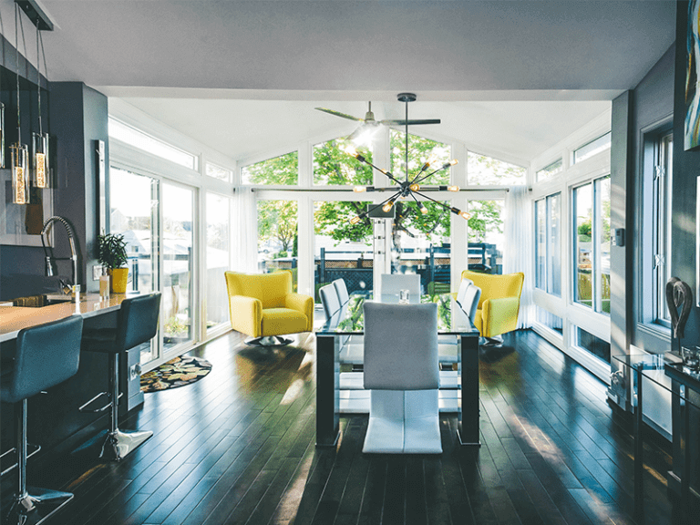 Modern, well-lit dining room featuring a blend of contemporary and cozy elements, highlighted by a stylish glass dining table, bright yellow armchairs, and a unique ceiling fan, with large windows providing ample natural light and a view of the outdoors.
