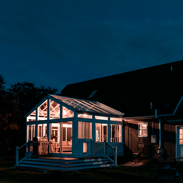 Two people sit at a desk working on a laptop inside a warmly lit, modern sunroom with glass ceilings and walls during twilight, creating a cozy, productive ambiance. A camera on a tripod and a cat tree are also visible, adding to the homely feel of the room.