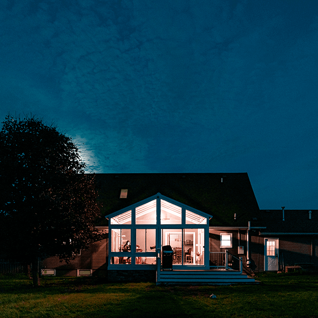 A cozy, well-lit sunroom with large windows is attached to a traditional dark-roofed house, captured during the twilight hours, creating a serene and inviting atmosphere.