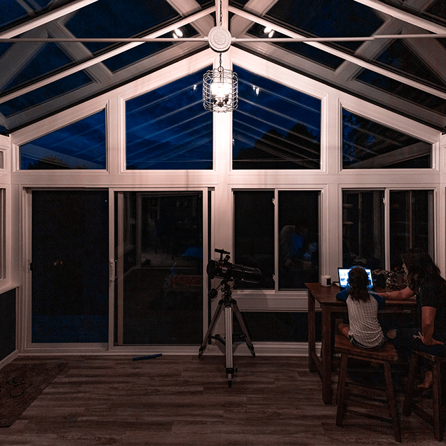 A cozy, illuminated sunroom with large glass windows on the back of a residential home under a night sky, featuring interior lights that warmly glow against the dark, cloud-covered moonlit backdrop. Two figures can be glimpsed sitting comfortably inside, creating a serene and inviting atmosphere.