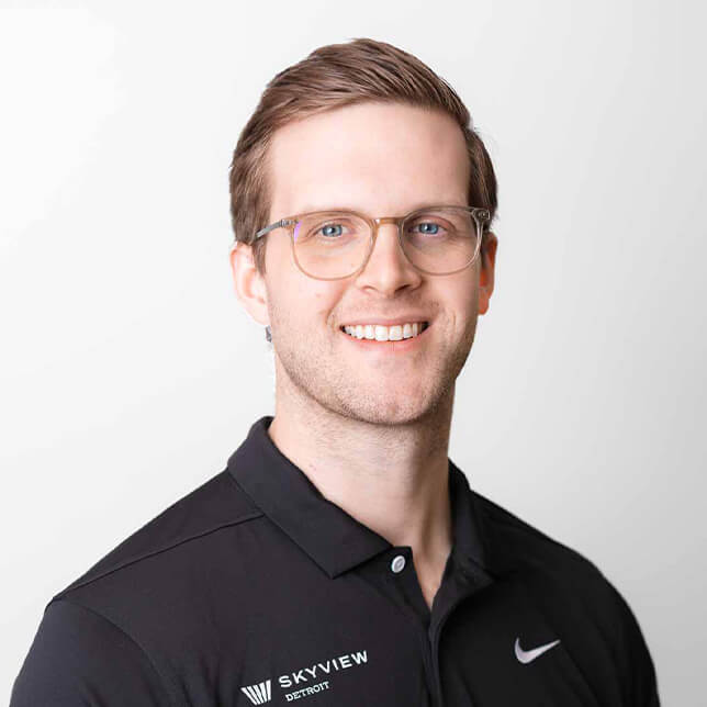 Portrait of a smiling man with light brown hair and round glasses, wearing a black Nike polo shirt with the 'Skyview Detroit' logo embroidered on it. He stands against a plain white background, presenting a friendly and professional appearance.