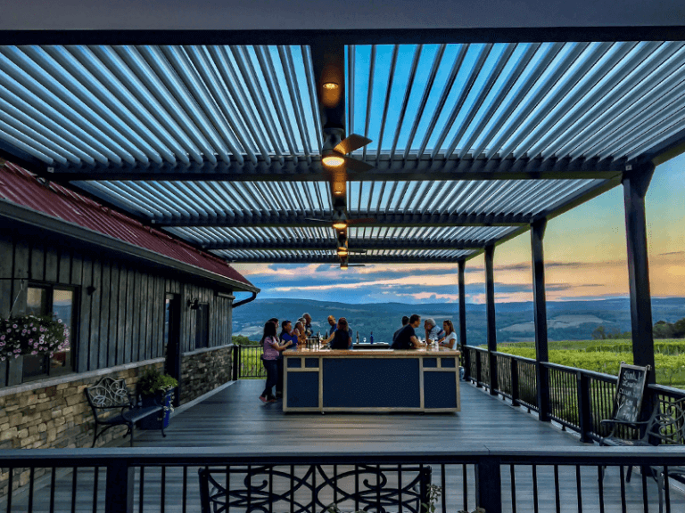 People enjoy a serene evening at an outdoor bar under a modern metal pergola, overlooking a vast landscape with mountains at sunset, creating a relaxing and picturesque setting.
