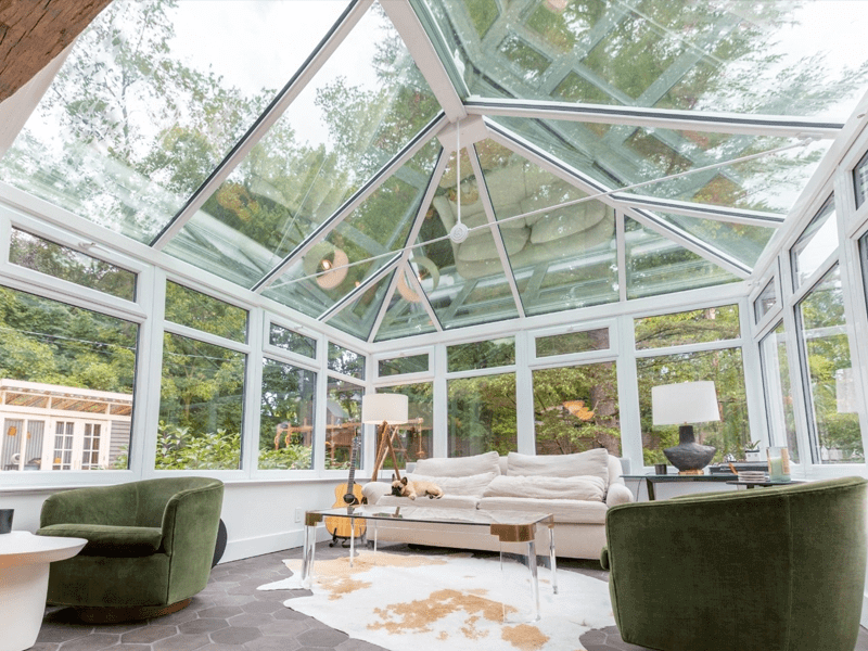 Luxurious sunroom with a high glass ceiling and full-length windows surrounded by lush greenery, featuring stylish furnishings including a plush green armchair and a cozy white sofa. The room's modern yet homey design showcases a unique blend of comfort and contemporary aesthetics, perfect for relaxation and enjoying natural views.
