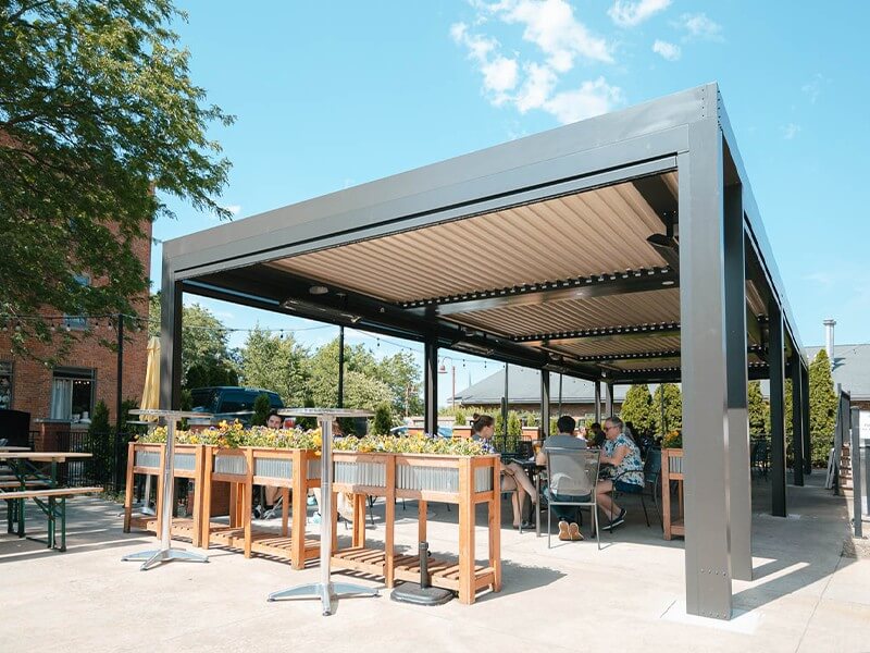 Outdoor dining area at a modern restaurant featuring a large metal pergola with retractable roof, wooden tables surrounded by lush planters, and people enjoying meals on a sunny day.