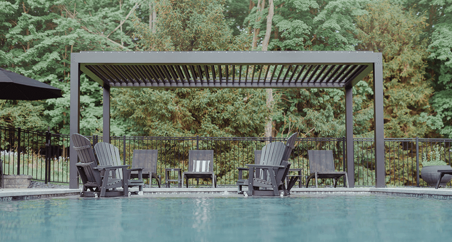 View of a spacious deck featuring modern outdoor furniture positioned under a pergola, overlooking a lush forest and distant water view under a bright sky.