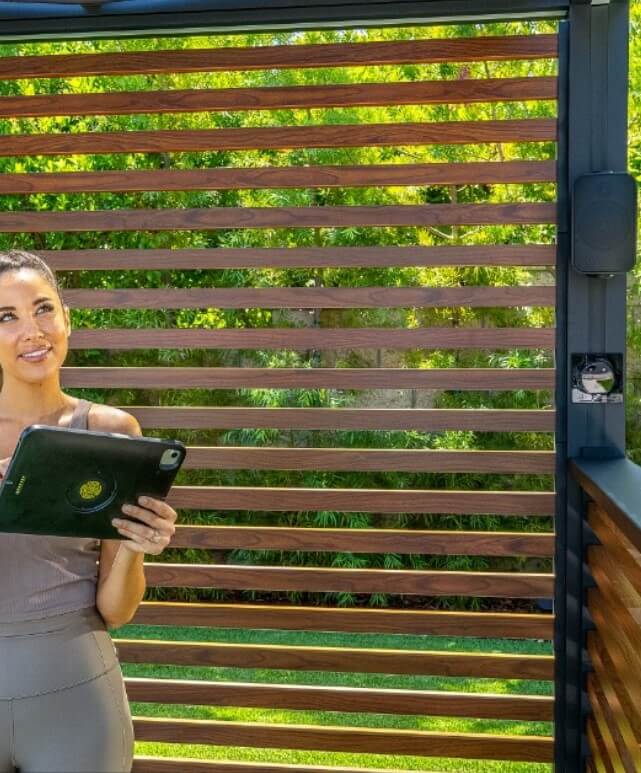A professional woman holding a tablet stands by a modern slatted wooden fence with lush greenery in the background, demonstrating technology in an outdoor setting.