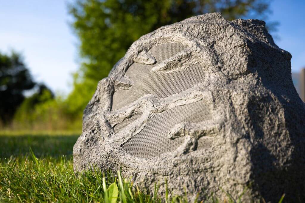 Close-up of a rock speaker disguised as a stone with dinosaur fossils on it, set against a vibrant green lawn and a blurred background of trees under clear skies. This outdoor speaker integrates seamlessly into garden settings, blending technology with natural aesthetics.