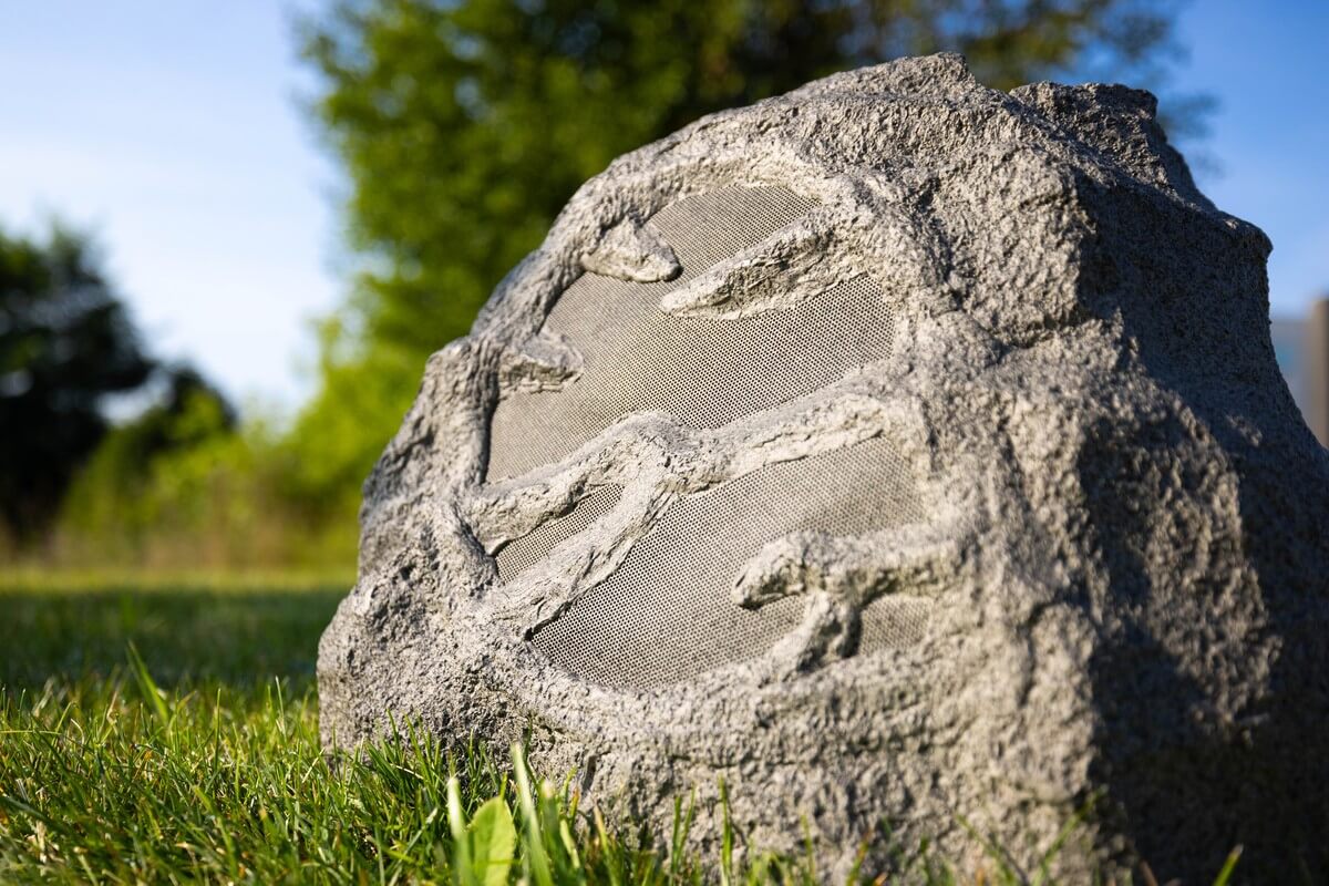 Close-up of a rock speaker disguised as a stone with dinosaur fossils on it, set against a vibrant green lawn and a blurred background of trees under clear skies. This outdoor speaker integrates seamlessly into garden settings, blending technology with natural aesthetics.