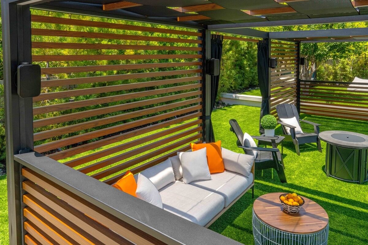 Modern outdoor living space featuring a stylish pergola with horizontal wooden slats, comfortable seating including a sofa with orange cushions and chairs, set in a lush green garden. Bright sunlight filters through the slats, casting shadows on the vibrant, manicured turf.