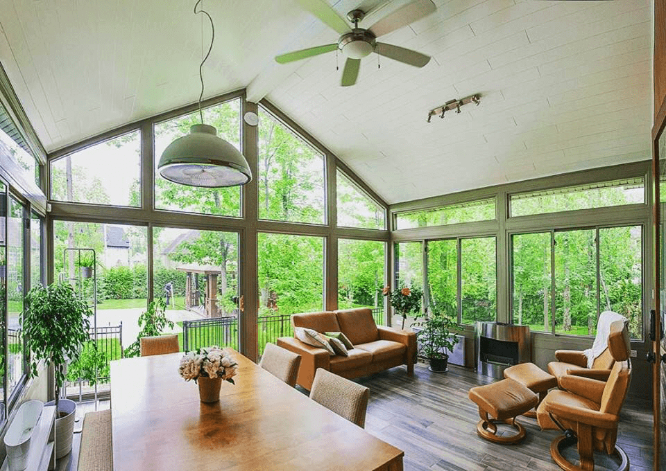 A cozy, well-lit sunroom with clear glass ceilings and ample windows showcasing a lush green backyard. The room features a plush beige sofa occupied by a sleeping dog, a classical guitar leaning against a side, and a stylish table lamp on a dark table, creating a serene indoor space.