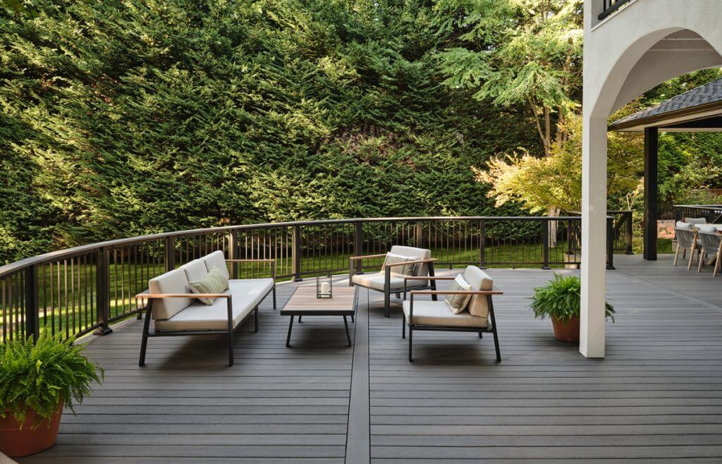 A serene outdoor living space featuring modern furniture with neutral cushions on a dark wooden deck, surrounded by lush greenery and a metal railing. This inviting area is ideal for relaxation or entertaining guests in a natural setting.