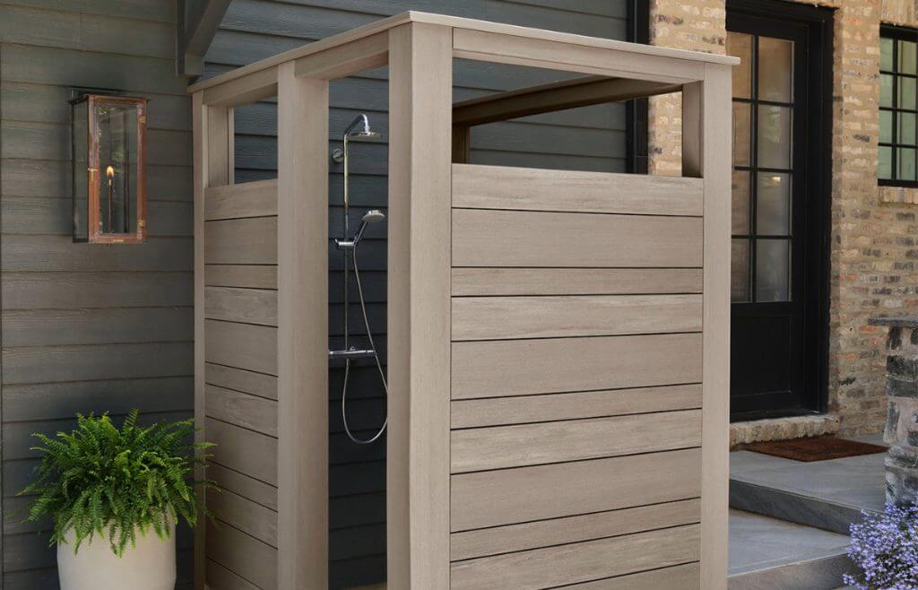 An outdoor shower enclosed with modern wooden privacy panels, set against a contemporary home exterior with dark siding and a rustic lantern, showcasing a blend of functionality and aesthetic design.