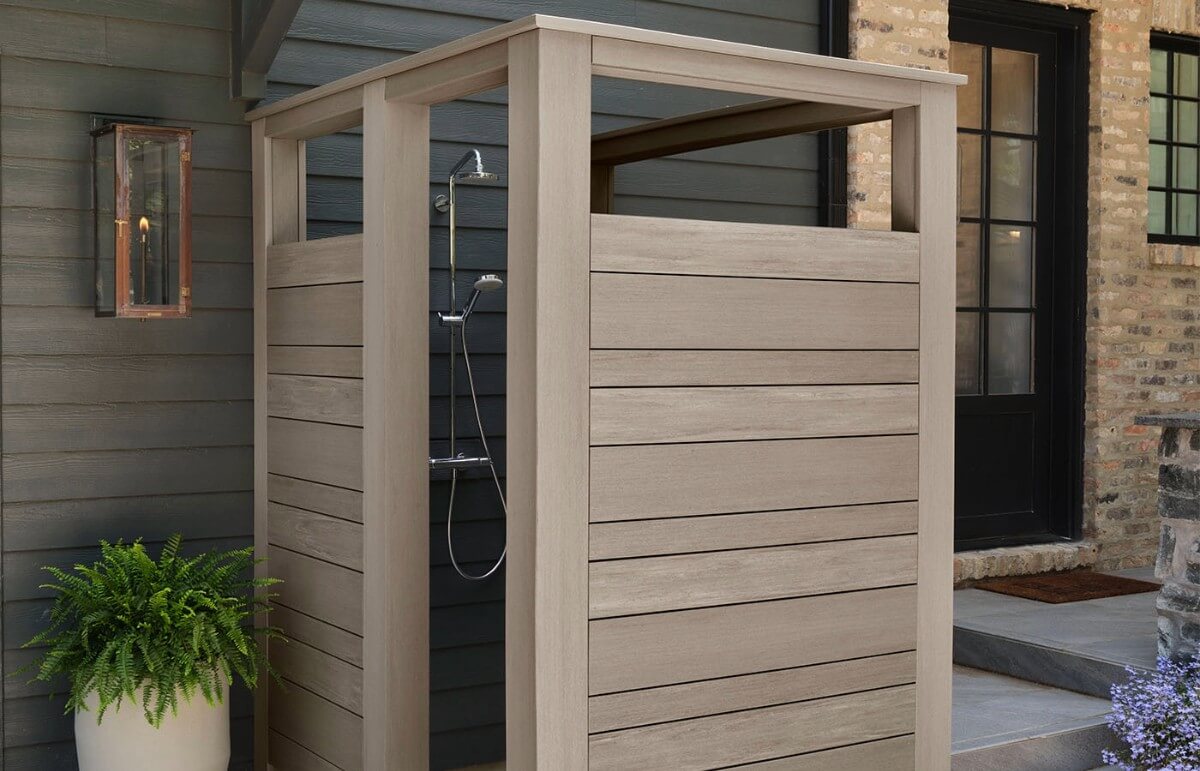 An outdoor shower enclosed with modern wooden privacy panels, set against a contemporary home exterior with dark siding and a rustic lantern, showcasing a blend of functionality and aesthetic design.