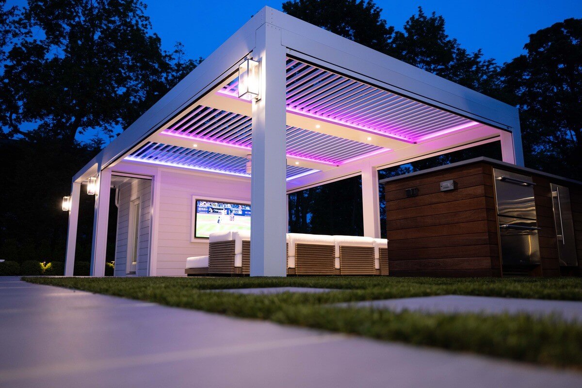 An elegant outdoor seating area under a modern pergola illuminated by pink and purple LED lights during twilight, featuring comfortable seating and a large screen TV, surrounded by a well-manicured lawn.
