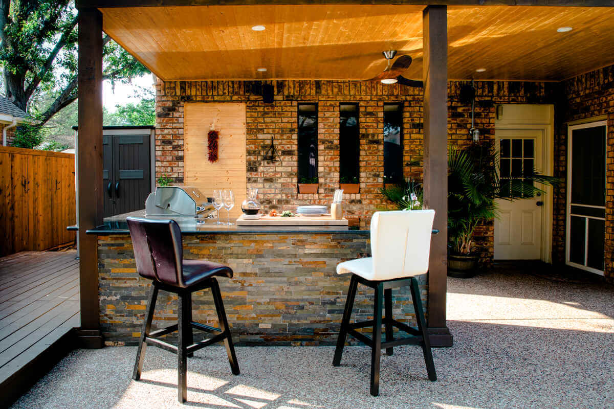outdoor kitchen built against a brick house with a covered roof, stone bar island, built-in grill, and bar seating