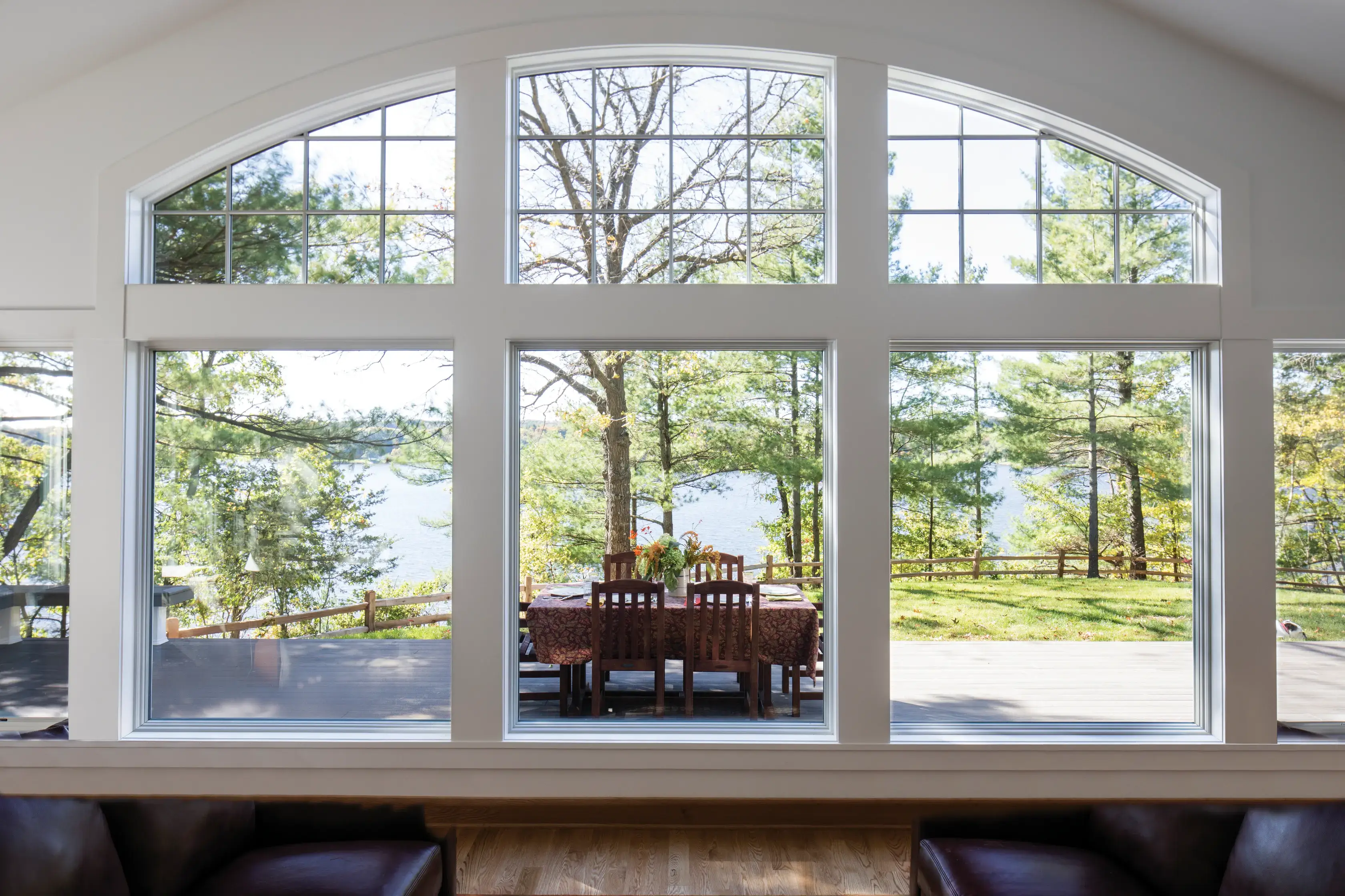Large picture window with white frames overlooking a lakeside backyard with trees, wooden deck, and outdoor dining table.