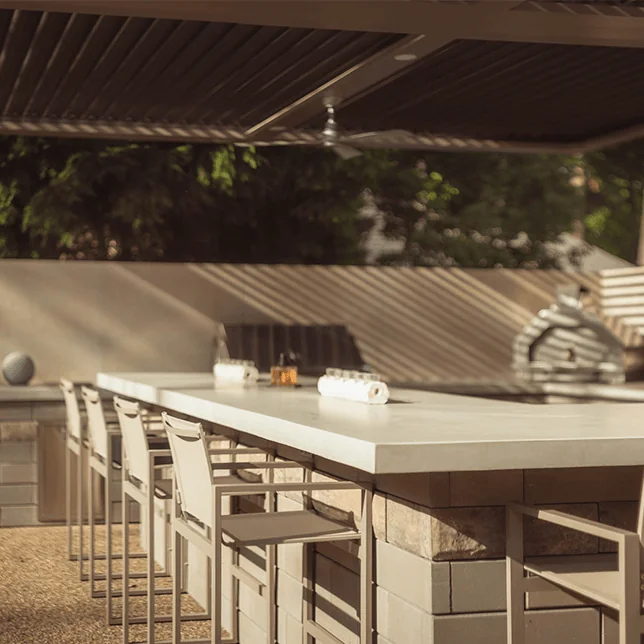 Elegant outdoor kitchen and bar seating area under a shaded pergola, featuring a modern stone bar counter lined with high stools, and completed with sleek, ambient lighting.
