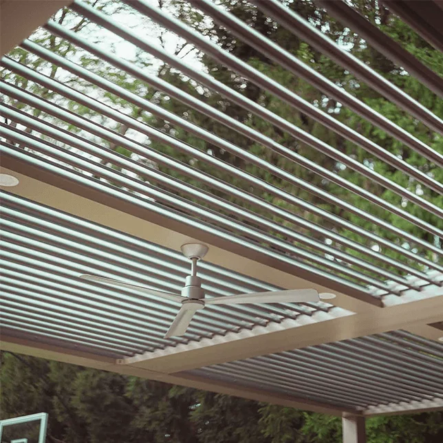 Modern outdoor patio area featuring a ceiling fan mounted on a slatted wooden canopy with green trees visible through the slats, suggesting a blend of technology and nature in home design.