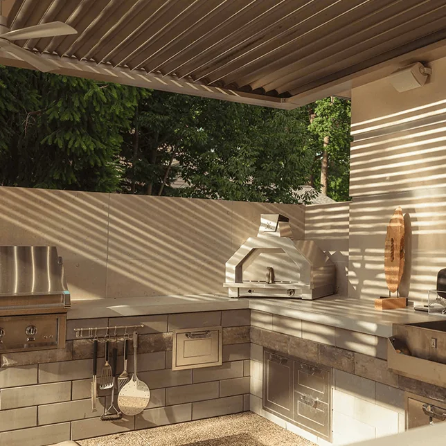 Outdoor kitchen area featuring built-in stainless steel appliances and a pizza oven under a pergola, illuminated by natural sunlight filtering through the slats, set against a backdrop of lush green trees.