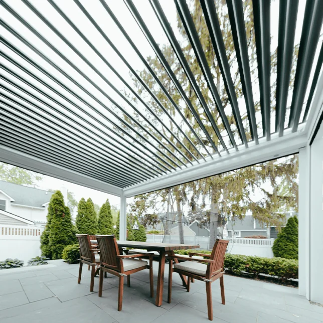 A cozy outdoor patio area with modern pergola cover, wooden dining table, and benches facing a well-manicured garden with tall conical trees and a view of neighboring houses. The space exemplifies a serene suburban backyard ideal for dining and relaxation.