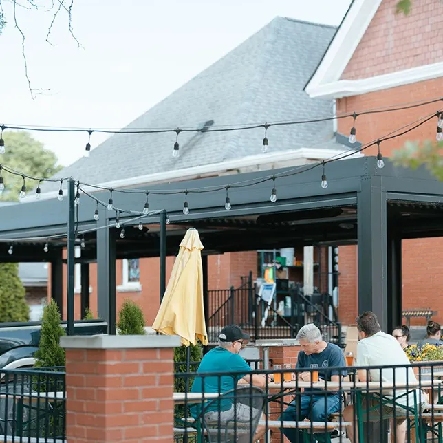 People dining at an outdoor restaurant patio under string lights, with a yellow umbrella and a view of a brick building in the background.