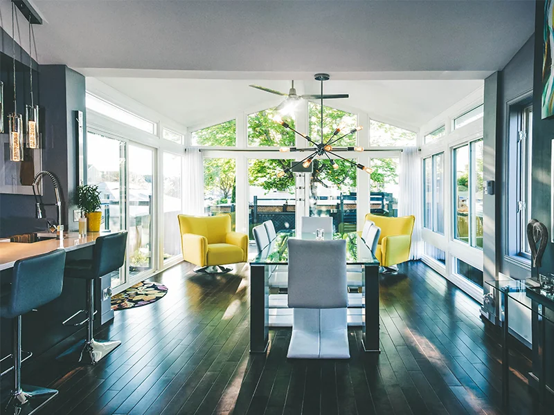 Modern, well-lit dining room featuring a blend of contemporary and cozy elements, highlighted by a stylish glass dining table, bright yellow armchairs, and a unique ceiling fan, with large windows providing ample natural light and a view of the outdoors.
