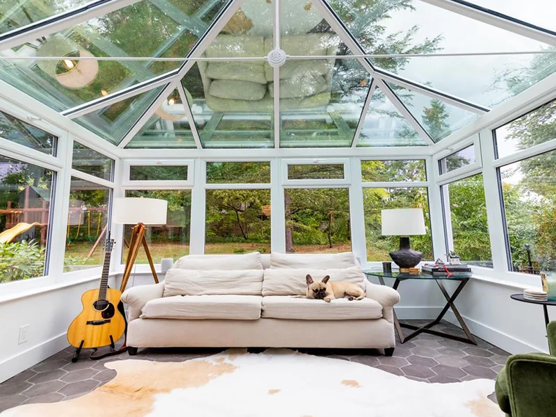 A cozy and well-lit sunroom featuring a large, cushioned sofa with a sleeping dog, surrounded by a lush garden view through glass paneled walls and ceiling, complemented by a classical guitar and elegant decor.