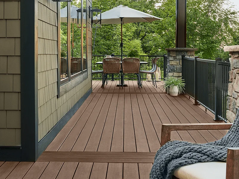 A cozy outdoor patio area featuring a wooden deck with two rattan chairs under a white umbrella, framed by lush green trees and a stylish boundary wall made of stone and metal railings. The scene also includes a large knitted throw on a bench, enhancing the inviting atmosphere of this serene outside living space.