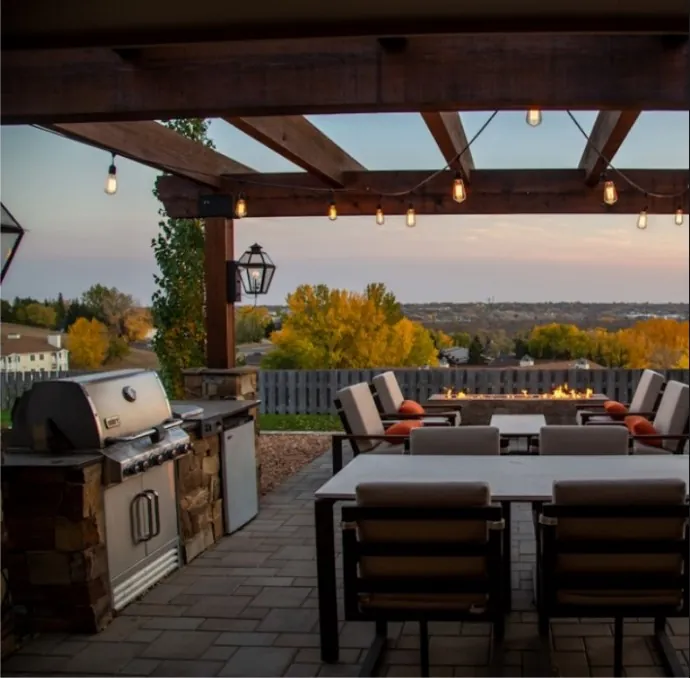 An inviting outdoor patio featuring modern furniture and a built-in barbecue grill, set under a wooden pergola adorned with hanging lights, overlooking a scenic landscape during a serene sunset.